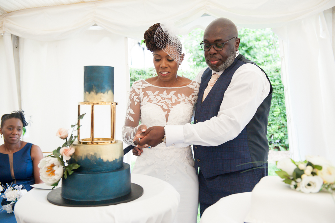 A couple cutting a teal and gold wedding cake in a marquee at a Nigerian wedding reception in Hertfordshire