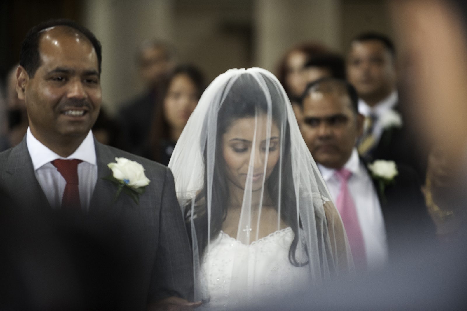 The bride behind her veil beside her father in the church aisle at an Asian wedding in London
