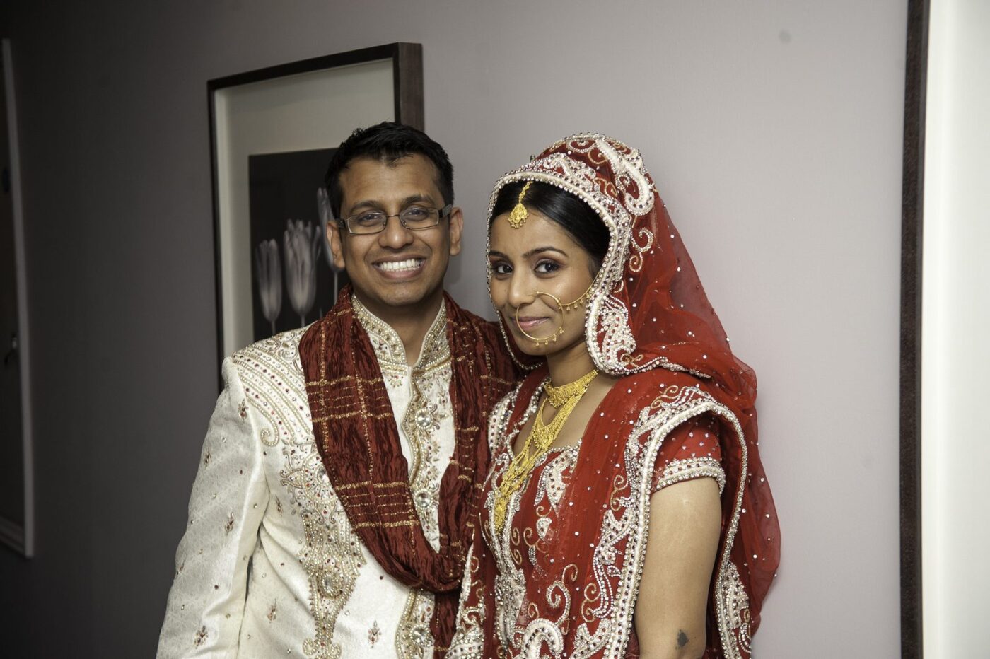 The bride in a red and gold lehenga with a dupatta over her head, a nath and a maang tikka, beside the groom in a cream sherwani