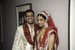 The bride in a red and gold lehenga with a dupatta over her head, a nath and a maang tikka, beside the groom in a cream sherwani