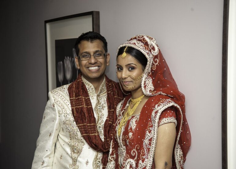 The bride in a red and gold lehenga with a dupatta over her head, a nath and a maang tikka, beside the groom in a cream sherwani