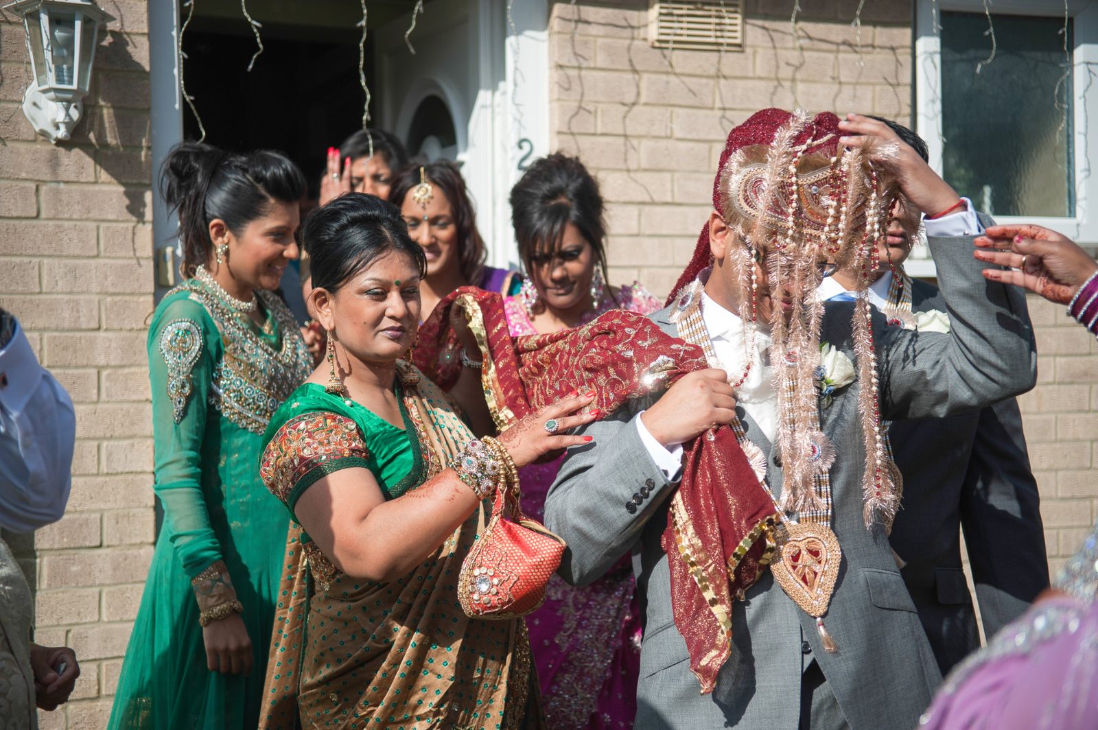 The groom leaving the family home with the sehra strands covering his face, family adjusting his shawl at an Indian wedding