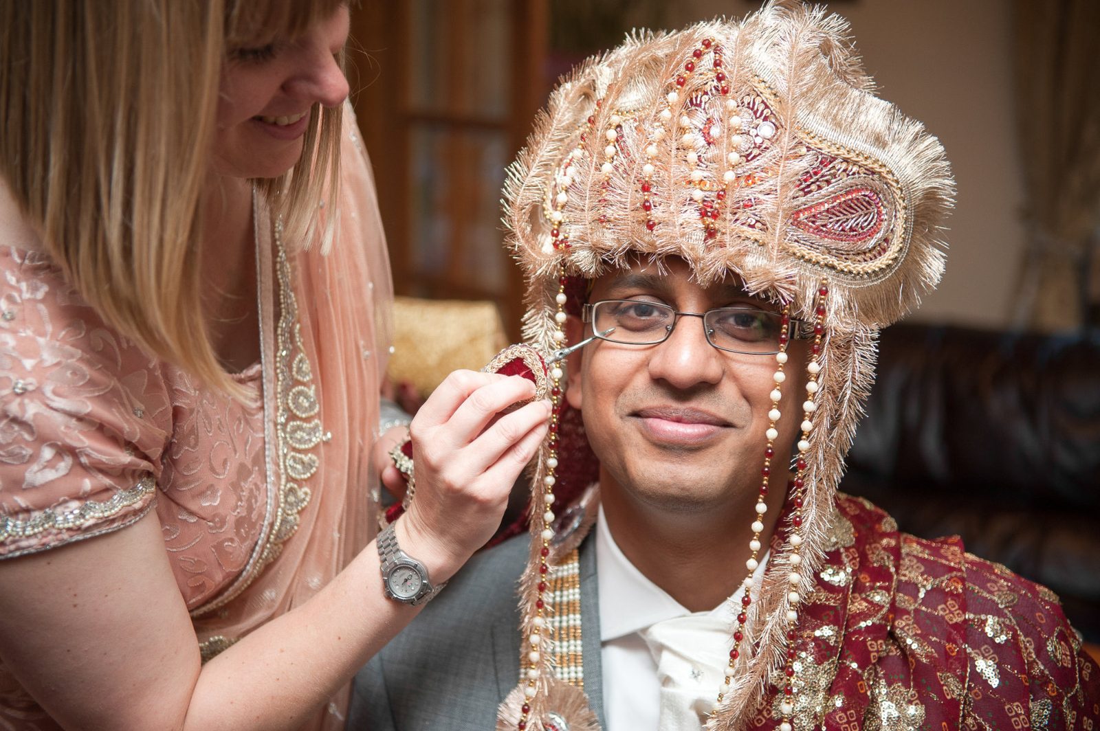 The groom wearing a sehra, the beaded and pearl fringed headdress tied over a turban base, being fitted at home before an Asian wedding in London