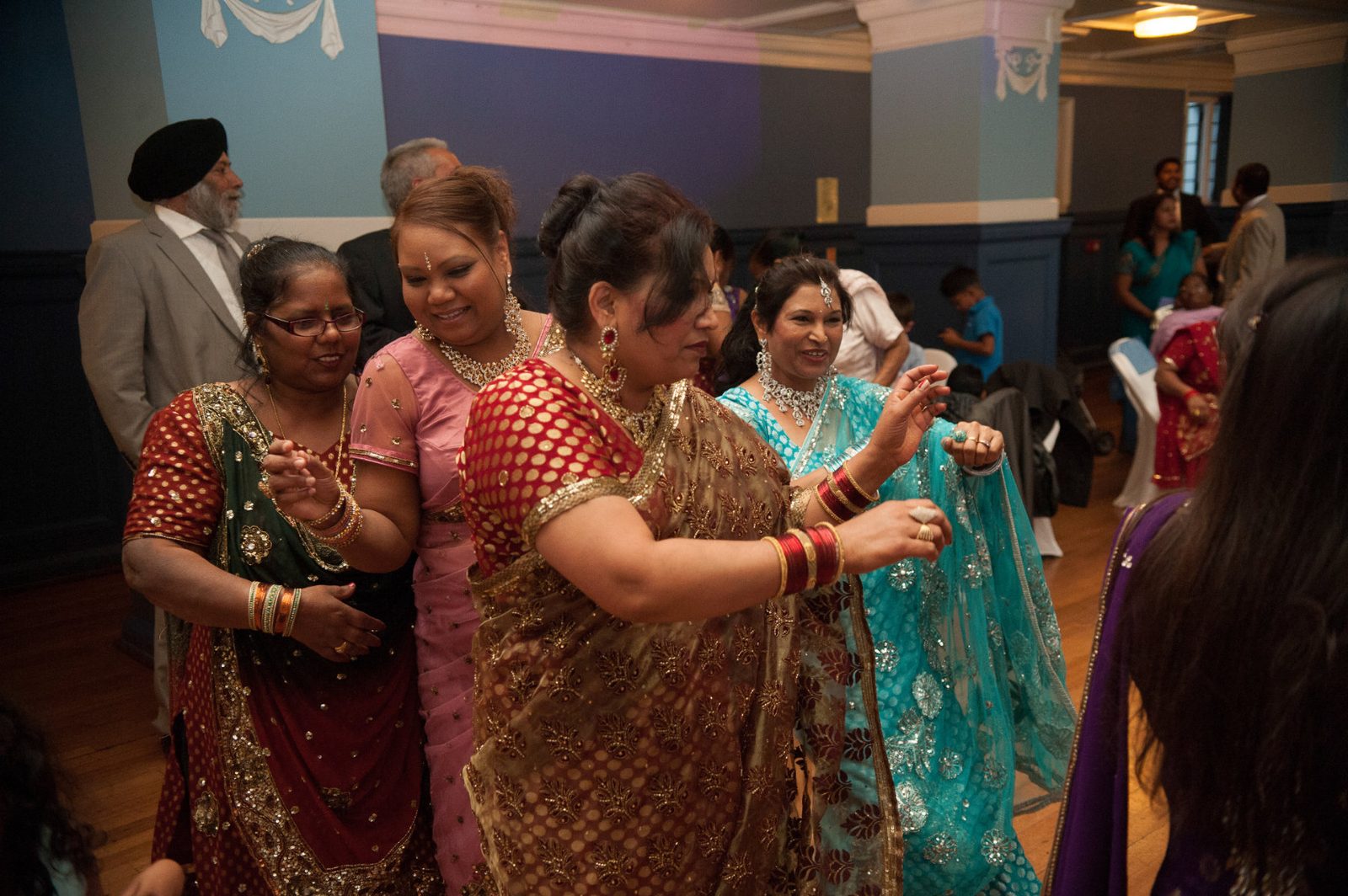 Women in saris and lehengas dancing together with red and gold chooda bangles at an Asian wedding reception
